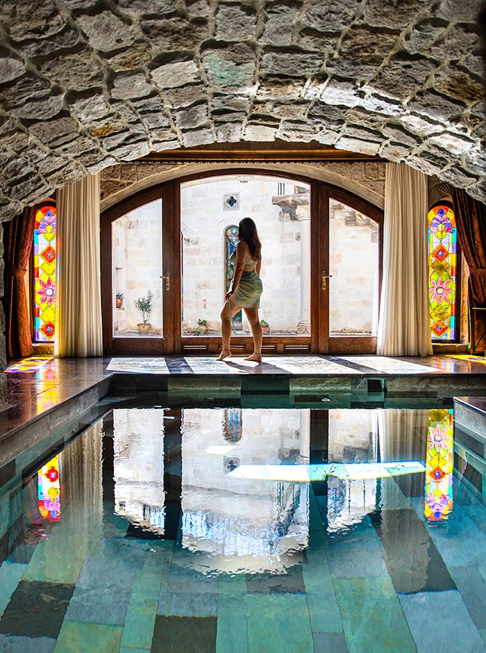 This image shows a woman standing by the edge of an indoor pool, framed by a stone-arched ceiling and stained-glass windows. The scene appears to be inside a luxurious cave-like setting, possibly a hotel suite, with reflections of the architecture and surroundings visible in the pool water. The bright stained-glass windows and stone architecture give it a historic, serene ambiance.