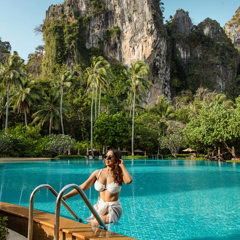 A woman in a white bikini and sunglasses stands at the edge of a large, inviting pool, surrounded by lush tropical vegetation and towering limestone cliffs. Palm trees and dense greenery create a serene, exotic atmosphere, with the cliffs providing a dramatic backdrop against a clear blue sky. The scene exudes relaxation and natural beauty.






