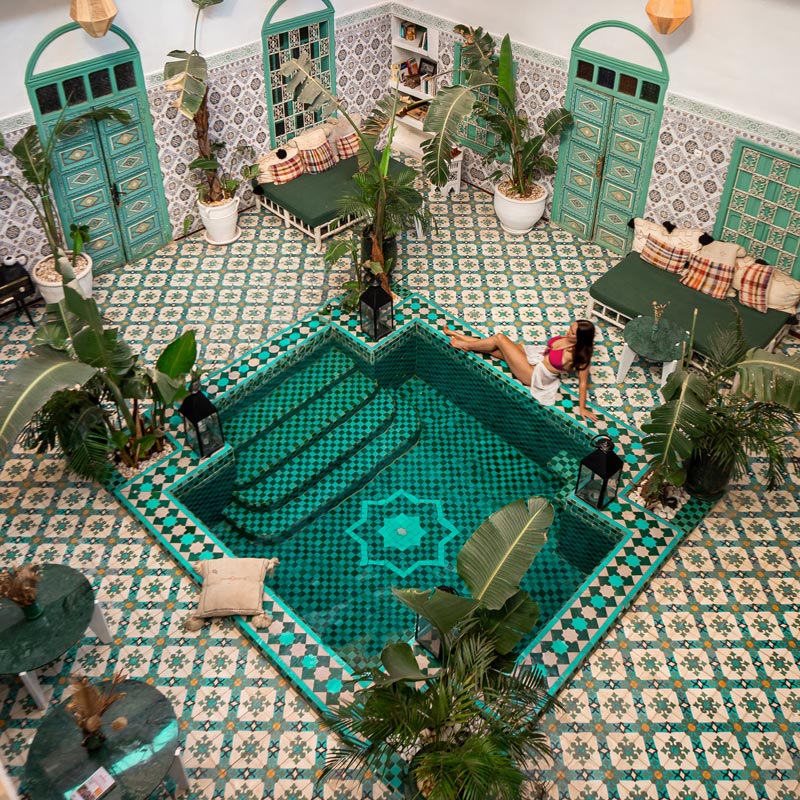 A vibrant courtyard featuring intricate green and white tiles surrounds a small, square pool with steps. A woman in a pink bikini and white towel sits by the pool's edge. The space is decorated with lush plants, green doors, and cozy seating areas adorned with cushions.






