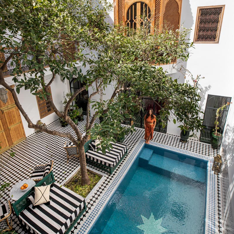 An ornate courtyard with black and white geometric tiles surrounds a rectangular pool. A woman in an orange dress stands beside the pool, with lush trees providing shade. The architecture features wooden doors and intricate latticework, creating a serene and inviting atmosphere.






