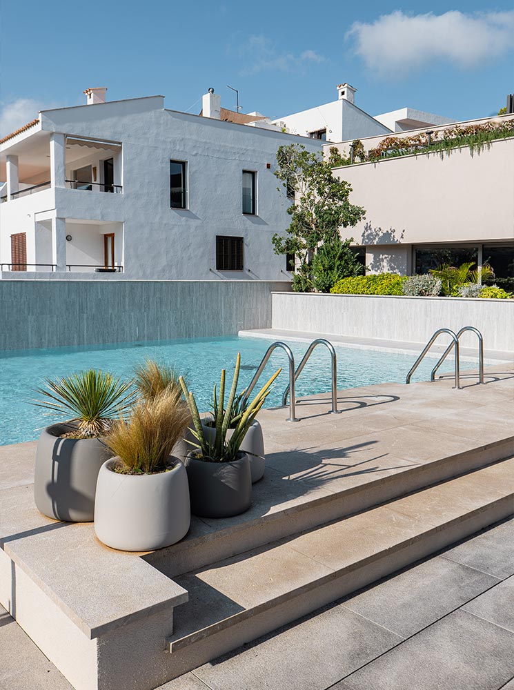 This image showcases a serene rooftop pool with steps leading into the water, accompanied by decorative potted plants featuring desert-style greenery. The background highlights whitewashed modern buildings and lush landscaping, creating a minimalist and sophisticated ambiance under a clear blue sky.