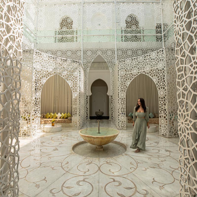 An elegant courtyard adorned with intricate white latticework features a central marble fountain. A woman in a flowing green dress walks through the space, which is surrounded by arched alcoves with plush seating and colorful pillows. The white and gold floor tiles complement the ornate, airy design, creating a luxurious and serene ambiance.






