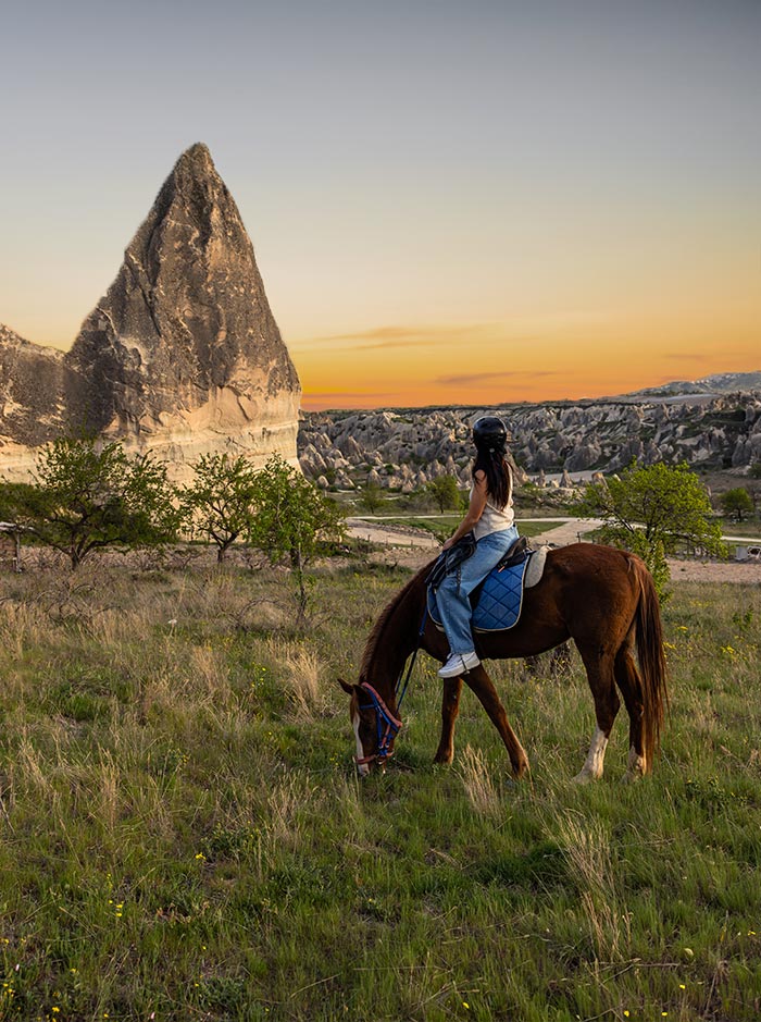 A woman wearing a helmet rides a brown horse on a grassy field at sunset, with a distinctive tall rock formation and the rocky landscape of Cappadocia, Turkey, in the background
