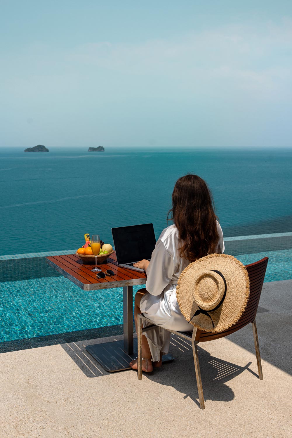 A woman wearing a white robe sits at a table by an infinity pool overlooking the ocean, working on a laptop. A straw hat rests on the back of her chair, and a bowl of fresh fruit and a glass of juice are on the table, suggesting a relaxing yet productive setting.