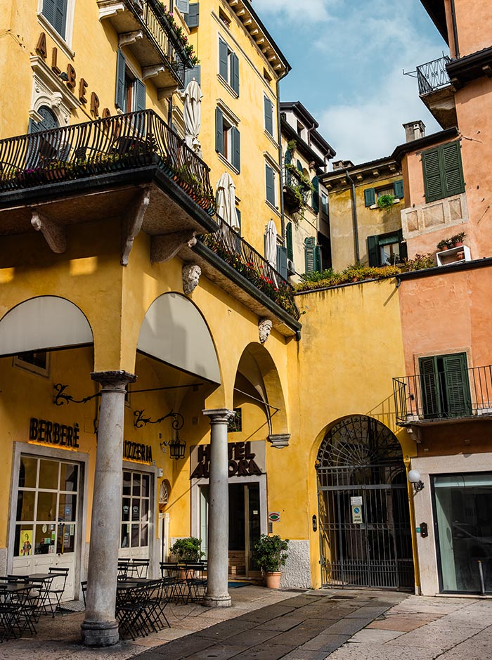 A charming street corner in Verona, featuring a yellow building with arched entrances and balconies adorned with plants. The ground floor houses Berberè Pizzeria, with its outdoor seating area set under the arches. The scene is bathed in soft daylight, highlighting the historic architecture and inviting atmosphere of this picturesque Italian city.