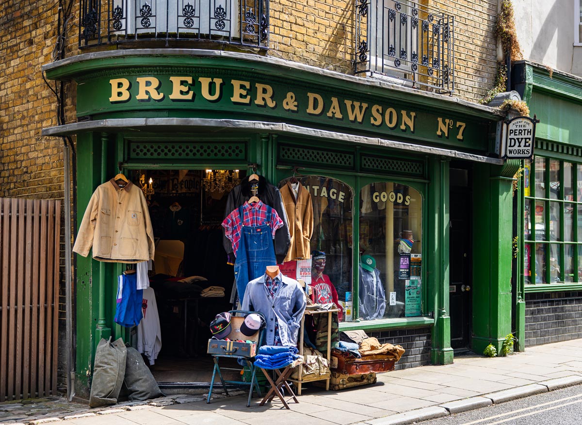 The storefront of Breuer & Dawson, a vintage goods shop, featuring a green facade with gold lettering. The display includes vintage clothing items such as overalls, jackets, and hats, arranged on mannequins and hangers outside the store. The entrance and windows showcase more items, inviting customers into the cozy, nostalgic atmosphere.