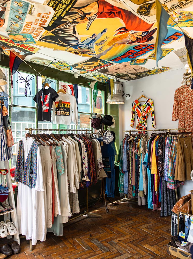 The interior of Breuer & Dawson vintage shop, featuring racks of colorful, eclectic clothing. The ceiling is decorated with vintage posters, adding to the retro ambiance. The room is filled with various garments, including dresses, shirts, and jackets, reflecting a diverse range of styles and eras. Bright light streams through the windows, highlighting the vibrant and nostalgic atmosphere of the shop.