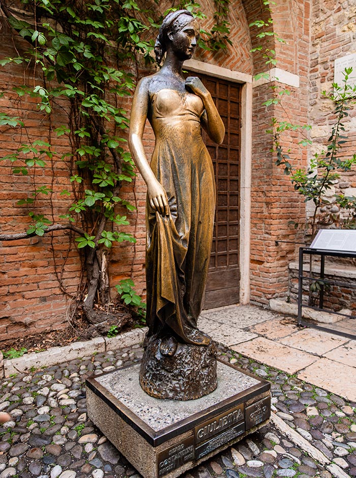 A bronze statue of Juliet (Giulietta) from Shakespeare's "Romeo and Juliet" stands in the courtyard of Casa di Giulietta in Verona. The statue depicts Juliet in a flowing dress, with one hand resting on her chest and the other holding her dress. The backdrop features brick walls with climbing vines, adding to the historic and romantic ambiance of the setting.