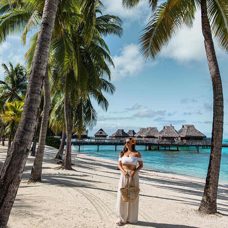 A woman in a white outfit with a straw bag and a flower in her hair strolls along a pristine sandy beach lined with tall palm trees. In the background, overwater bungalows at Conrad Bora Bora Nui extend over the turquoise lagoon. The tropical scenery, with clear blue skies and lush greenery, creates a perfect island paradise setting.






