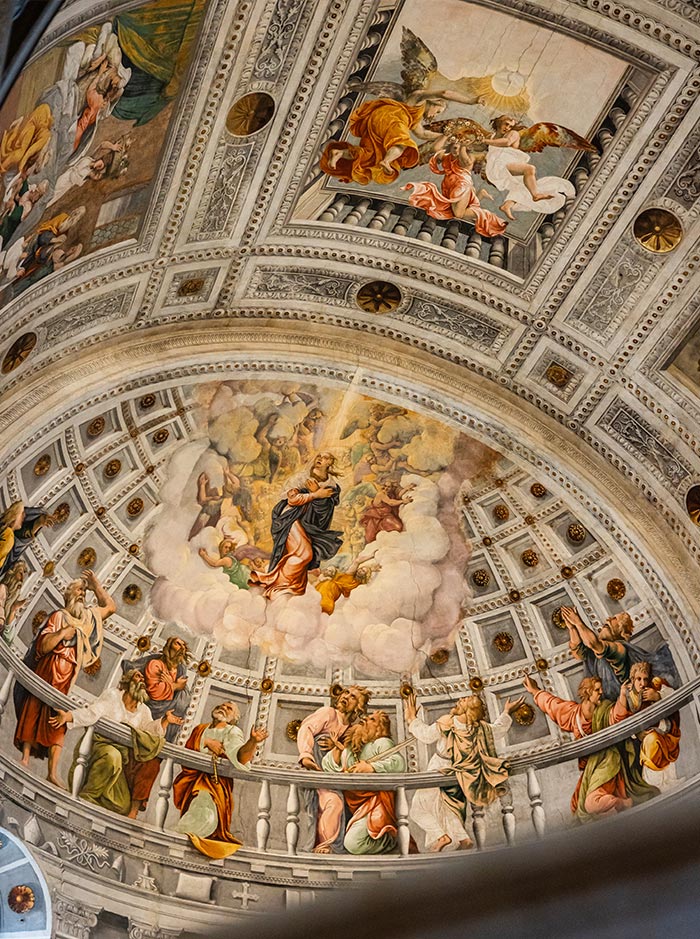 A stunning fresco adorns the ceiling of the Duomo di Verona, depicting religious figures and scenes in vibrant colors. The intricate artwork features heavenly figures and apostles, set against a detailed architectural backdrop with ornate patterns and a celestial theme. The craftsmanship and artistic detail highlight the historical and spiritual significance of this majestic cathedral.