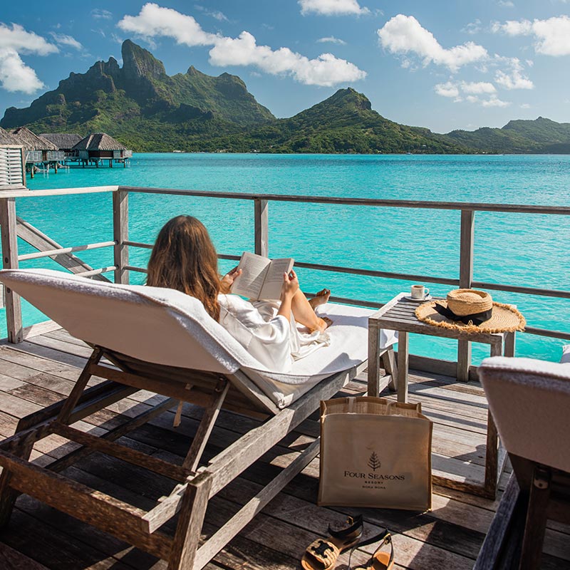 A woman lounges on a sunbed on a wooden deck overlooking the turquoise waters of Bora Bora at the Four Seasons Resort. She is reading a book, with a table beside her holding a coffee cup and a straw hat. The stunning backdrop features overwater bungalows and the lush, jagged peaks of Bora Bora, set against a bright blue sky with fluffy white clouds.







