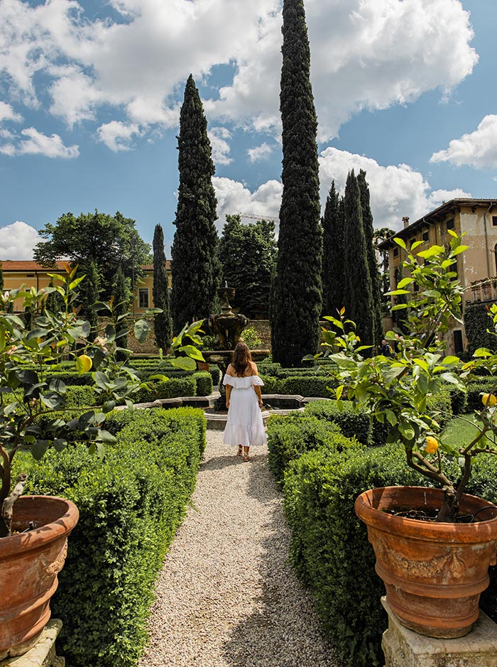 A woman in a white dress strolls through the manicured paths of Giardino Giusti, a historic garden in Verona. The scene features tall cypress trees, neatly trimmed hedges, and terracotta pots with lemon trees, all under a bright blue sky with fluffy clouds. The tranquil atmosphere is enhanced by the presence of a classical fountain at the center of the garden.