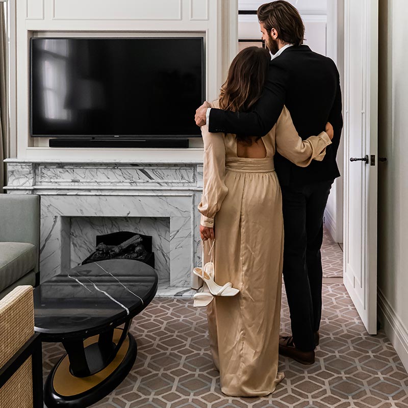 A couple dressed elegantly, with the woman holding her shoes, stands arm in arm facing a television and marble fireplace in a luxurious room at the Great Scotland Yard Hotel, part of Hyatt. The room features a stylish coffee table and patterned carpet.






