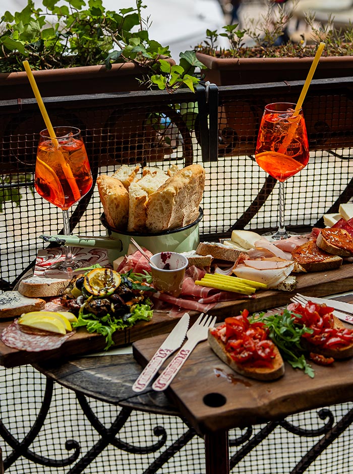 A colorful and appetizing spread of Italian antipasti is set on a table at La Prosciutteria in Verona. The meal includes various cured meats, cheeses, bruschetta with tomatoes and arugula, grilled vegetables, and a basket of fresh bread. Two glasses of Aperol Spritz add a vibrant touch to the scene, which is complemented by potted plants on a wrought-iron balcony railing. The setting is perfect for a leisurely meal, enjoying the flavors of Italy.