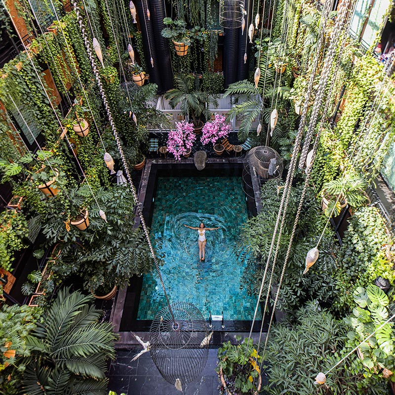 An indoor tropical oasis at Manon Les Suites in Copenhagen, featuring a serene pool surrounded by lush greenery and hanging plants. A woman in a white swimsuit floats peacefully in the center of the pool. The vibrant setting is enhanced by decorative lanterns and cascading foliage, creating a tranquil and exotic atmosphere.






