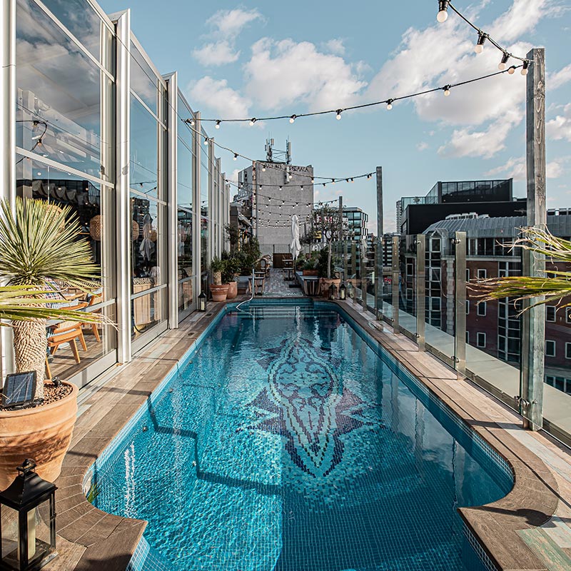 A rooftop pool at the Mondrian London Shoreditch, surrounded by glass barriers, potted plants, and string lights overhead. The pool features a mosaic design, and the view includes surrounding buildings and a clear sky with scattered clouds.






