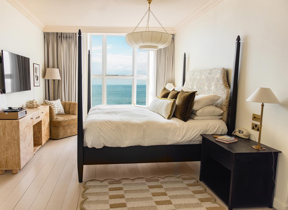 A well-lit, elegant bedroom with a black four-poster bed adorned with white and brown pillows, facing a large window showcasing a beautiful ocean view. The room features light-colored walls, a beige rug, a wooden side table, a cozy beige chair, and a TV mounted on the wall.