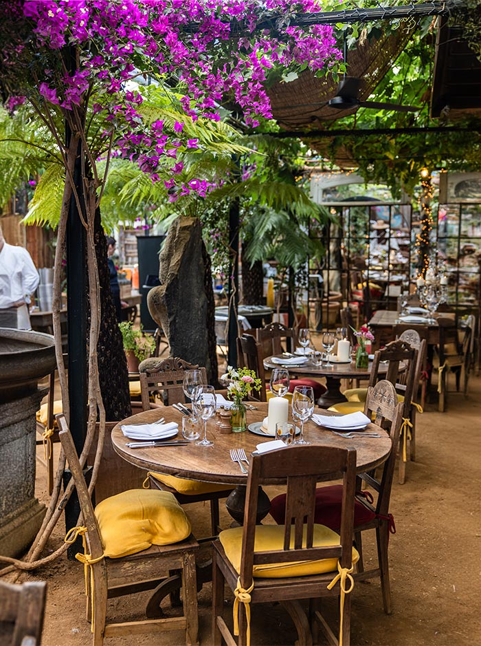 A cozy outdoor café at Petersham Nurseries in London, featuring wooden tables set with wine glasses, white napkins, and candles. The seating area is adorned with bright yellow cushions and surrounded by lush greenery and vibrant purple flowers hanging from above.