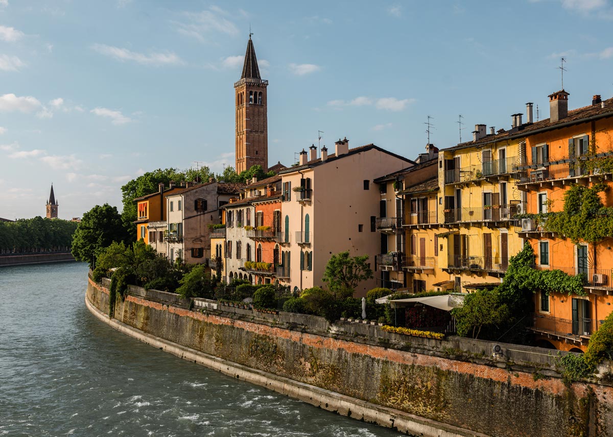 A picturesque view of the Adige River in Verona, lined with colorful buildings adorned with balconies and lush greenery. The scene is dominated by a tall bell tower rising above the rooftops, with another tower visible in the distance. The river winds gracefully through the city, bathed in the soft, golden light of late afternoon, creating a serene and charming atmosphere.