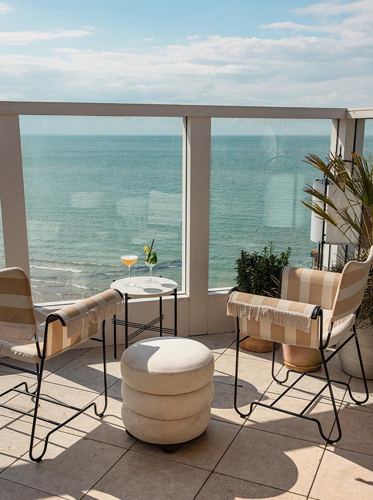 A serene rooftop bar setting at No. 42 Margate, featuring two striped armchairs and a round ottoman facing a stunning ocean view. A small table between the chairs holds a cocktail and a glass of wine, while potted plants add a touch of greenery to the cozy, sunlit space. The clear glass railing provides an unobstructed view of the sea and sky.