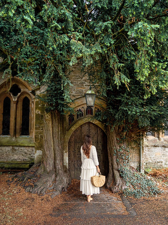 A woman in a flowing white dress stands before the arched wooden door of St. Edward's Church in the Cotswolds. The door is framed by ancient yew trees, creating a mystical and serene atmosphere. The historic stone building features arched windows and ivy-clad walls.