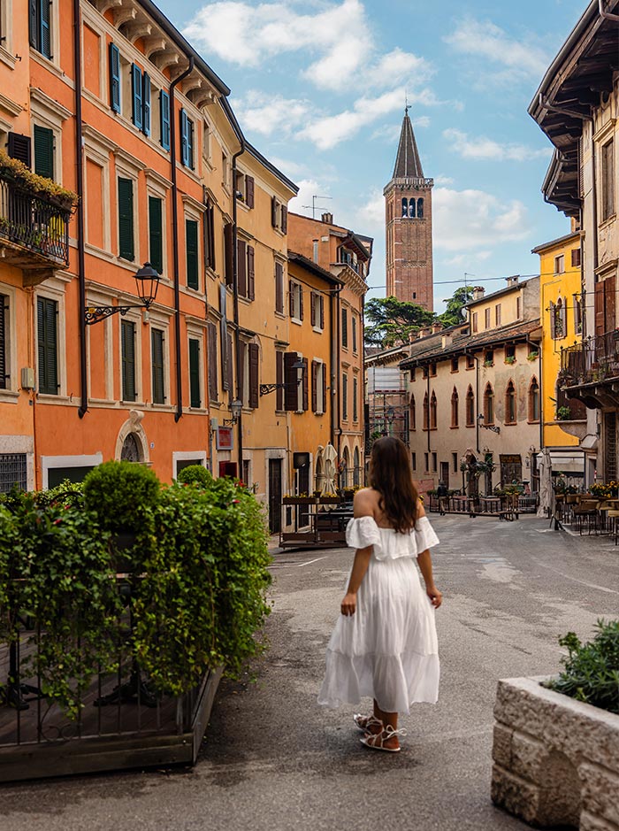 A woman in a white dress walks down a charming, narrow street in Verona, lined with colorful buildings and quaint balconies. The scene captures the city's historic ambiance with a bell tower rising in the background. The quiet street is adorned with greenery and outdoor seating, inviting a leisurely exploration of the picturesque Italian town.
