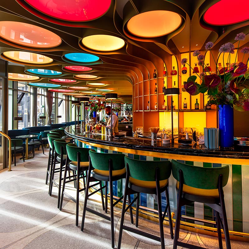 A vibrant bar at The BoTree, part of Preferred Hotels and Resorts, featuring colorful circular ceiling lights, a curved bar with high stools, and a variety of bottles displayed on illuminated shelves. The bar area includes lush floral arrangements and a bartender preparing drinks.






