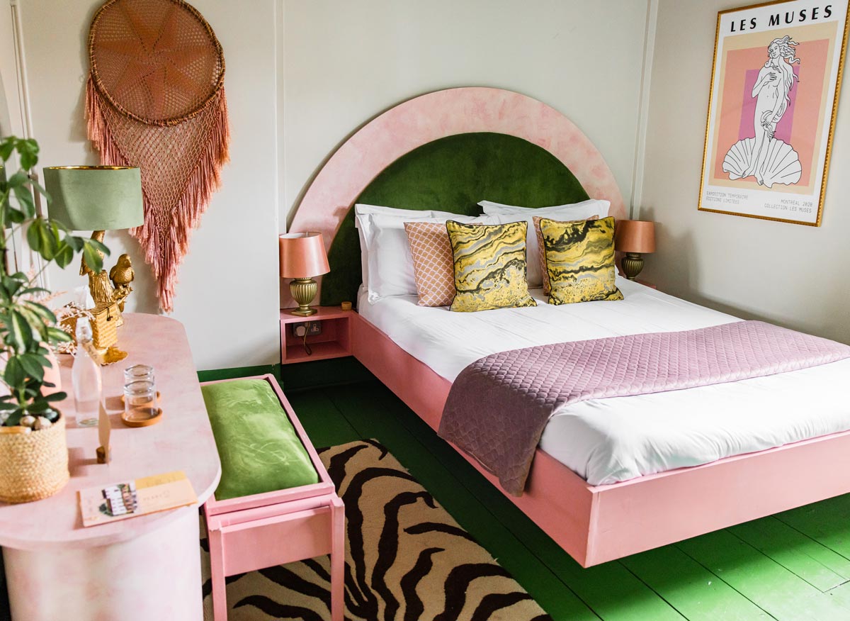 A vibrant and eclectic bedroom featuring a pink and green color scheme. The bed has a large pink and green headboard with patterned pillows and a purple throw blanket. The room includes a pink vanity with a green cushioned bench, a zebra print rug, and a large macramé wall hanging. A framed "Les Muses" poster and decorative items add artistic touches to the space.






