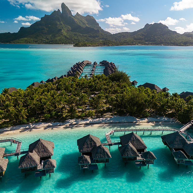 An aerial view of The St. Regis Bora Bora Resort, showcasing overwater bungalows extending into the clear turquoise waters. The resort is surrounded by lush palm trees and pristine beaches, with the dramatic peaks of Bora Bora in the background. The vibrant colors and stunning natural scenery create a picture-perfect tropical paradise.






