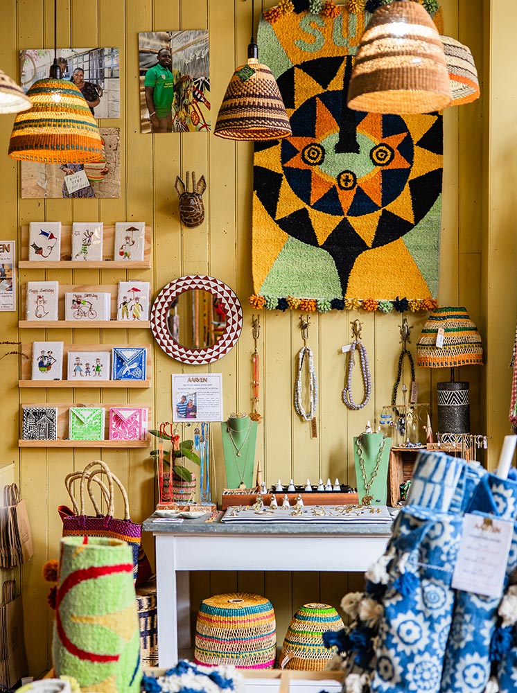 The interior of Aarven shop in Margate, showcasing a variety of handcrafted goods. The walls are adorned with colorful woven baskets, vibrant wall hangings, and jewelry displays. A central table features necklaces and other accessories, while shelves hold intricate cards and decorative items. The shop is brightly lit, creating a warm and inviting atmosphere filled with unique, artisanal products.