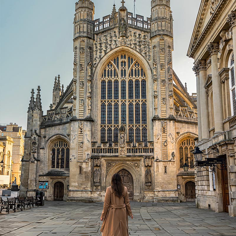 A woman in a long brown coat stands in front of the majestic Bath Abbey, an iconic Gothic cathedral with intricate architecture and large stained glass windows. The scene captures the grandeur of the abbey and the surrounding historic buildings, with a clear sky providing a perfect backdrop. The quiet, empty square emphasizes the abbey's impressive facade and serene atmosphere.







