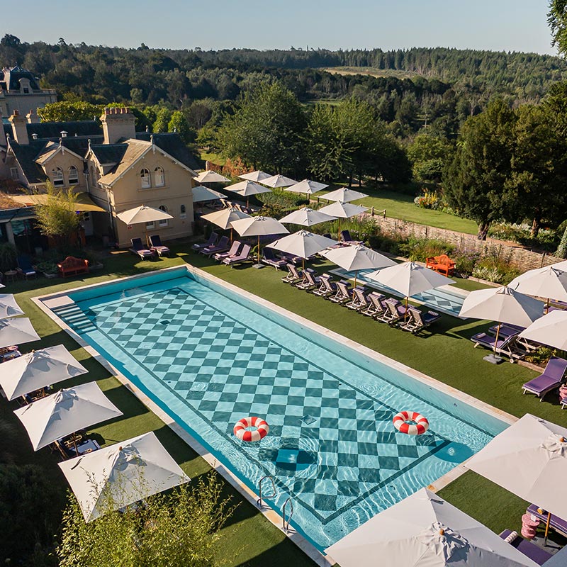 A luxurious outdoor pool area at Beaverbrook Hotel & Spa, featuring a large checkerboard-patterned pool surrounded by sun loungers with purple cushions and white umbrellas. The pool is set in a beautifully landscaped garden with lush greenery and a historic building in the background. The scene exudes relaxation and elegance, perfect for a tranquil getaway.






