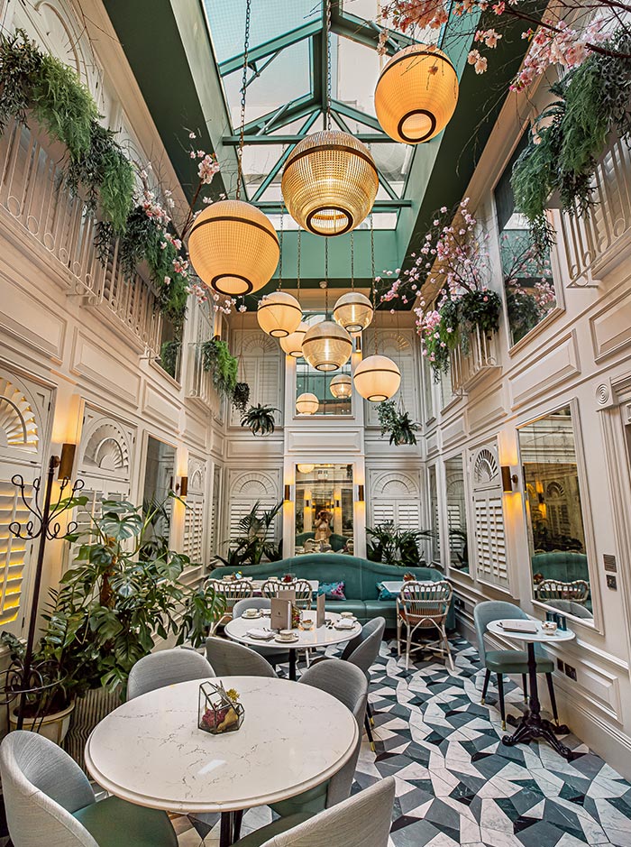 An elegant café at 100 Queen's Gate, London, featuring marble tables, plush chairs, and a cozy teal banquette. The space is decorated with hanging plants and cherry blossoms, with large globe lights hanging from a glass ceiling that lets in natural light. The floor is patterned with geometric tiles, adding to the chic and sophisticated ambiance.