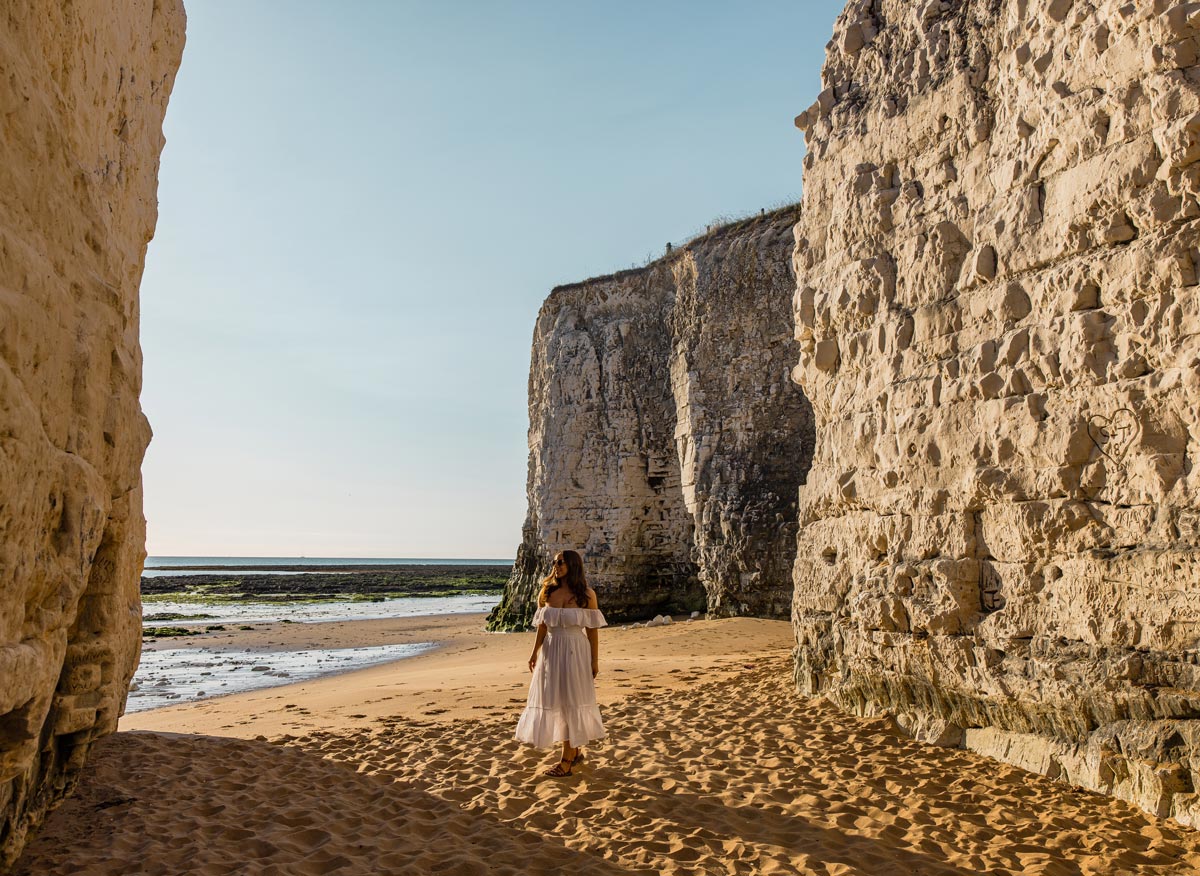 A woman in a white off-the-shoulder dress stands on a sandy beach between towering white chalk cliffs. The beach extends towards the calm sea in the background, with the sun casting a warm glow on the cliffs and sand. The scene is serene and picturesque, with the natural beauty of the coastal landscape on display.