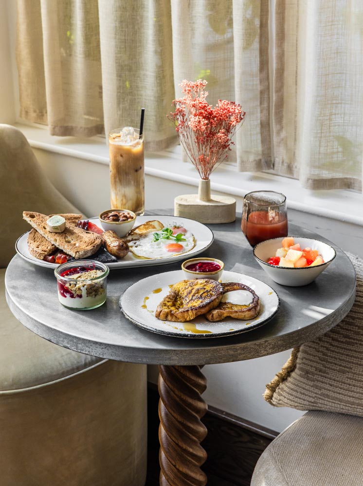 A charming breakfast setup at No. 42 Margate, featuring a variety of dishes on a stylish round table. The spread includes a plate of sunny-side-up eggs with toast and sausages, French toast with syrup and berry compote, a bowl of fresh fruit, and a glass of iced coffee. The table is decorated with a small vase of dried flowers, and the soft natural light filtering through the curtains creates a cozy and inviting atmosphere.