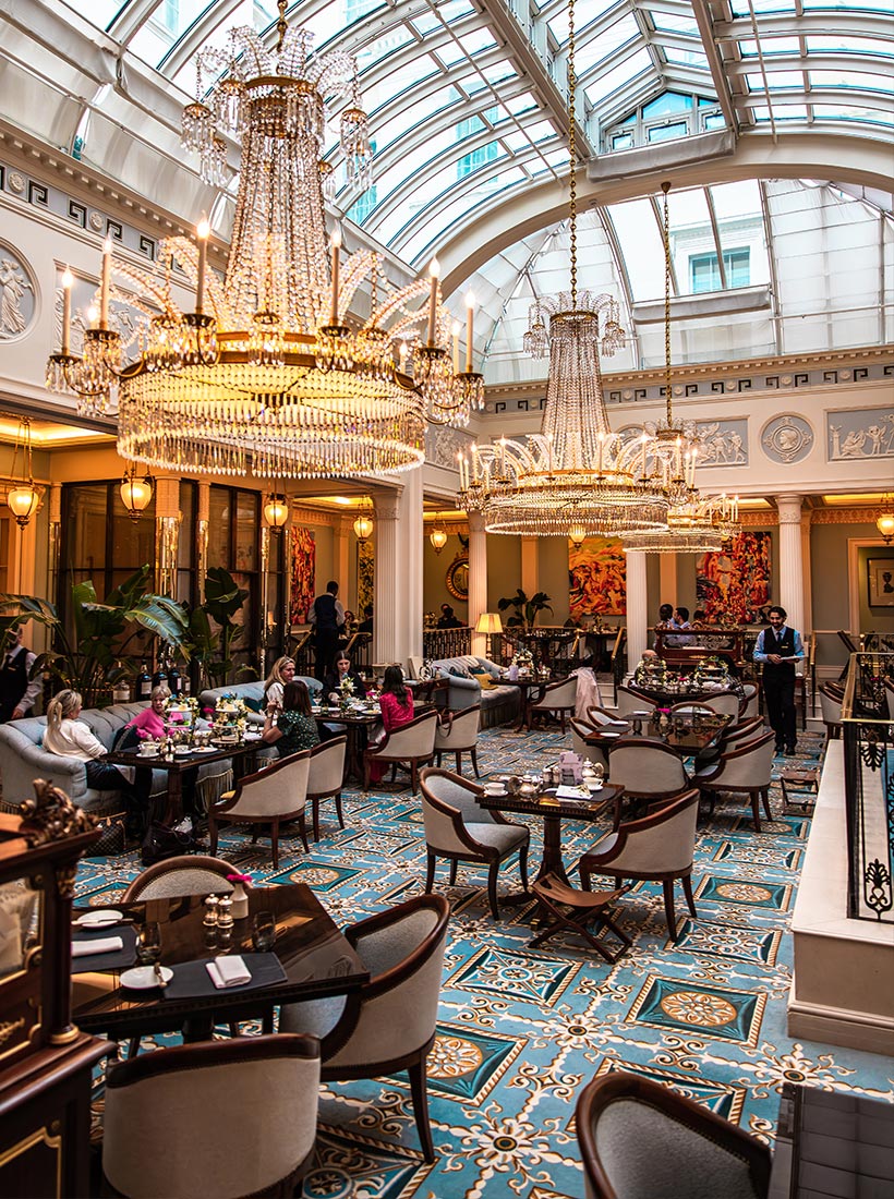 Large elegant tea room with a glass atrium ceiling and grand crystal chandeliers hanging above guests enjoying afternoon tea at small tables. People sit on upholstered chairs around tiered stands of pastries and sandwiches while a server walks through the bright ornate dining space. The setting features patterned blue tile flooring classical columns and warm lighting that creates a luxurious afternoon tea atmosphere.