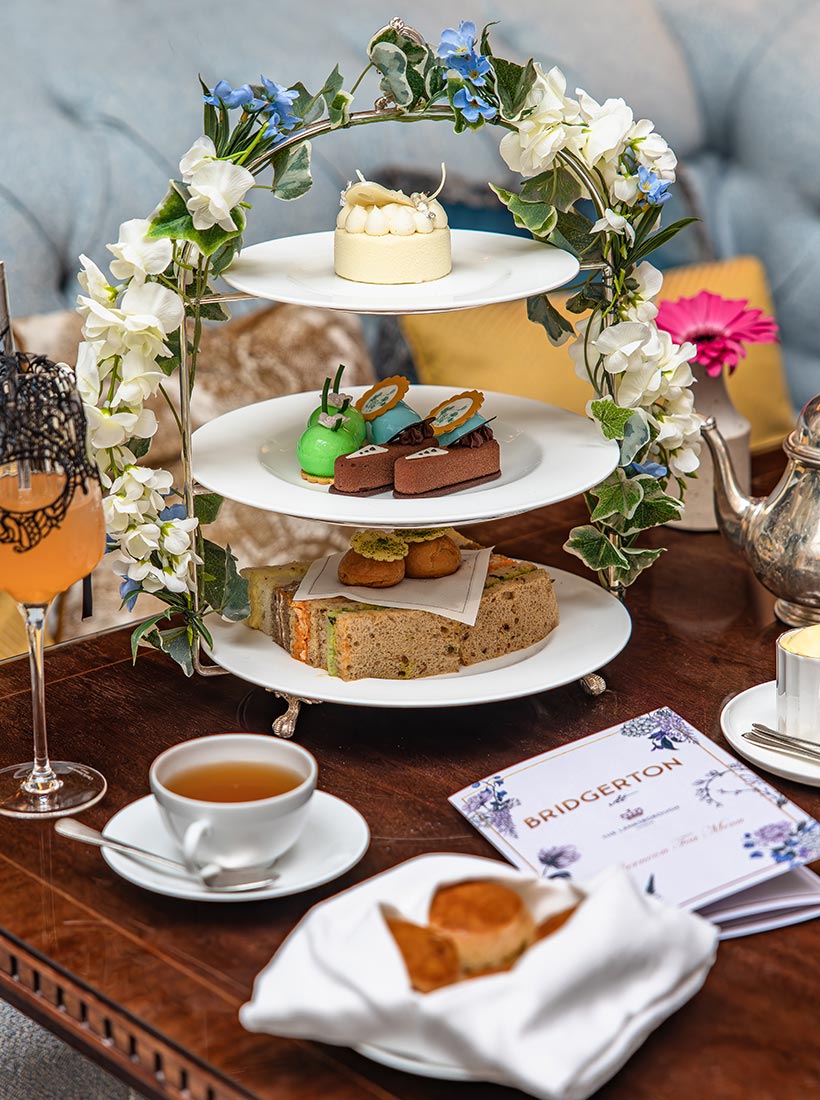 Three tier afternoon tea stand decorated with white flowers and blue blossoms holding small cakes pastries and finger sandwiches on a wooden table. A teacup a scone on a plate a cocktail and a silver teapot sit nearby with a card that reads Bridgerton suggesting a themed afternoon tea setting. The display creates an elegant garden inspired tea service presentation.