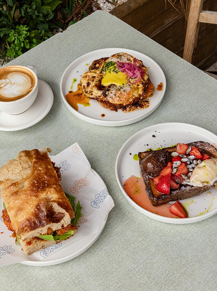 A delightful brunch setup at Forts in Margate, featuring a cappuccino, a savory sandwich, a dish of eggs with hollandaise sauce and pickled onions, and a plate of French toast topped with strawberries and cream. The dishes are artfully presented on a light green table, with a backdrop of greenery adding a fresh and inviting touch to the meal.