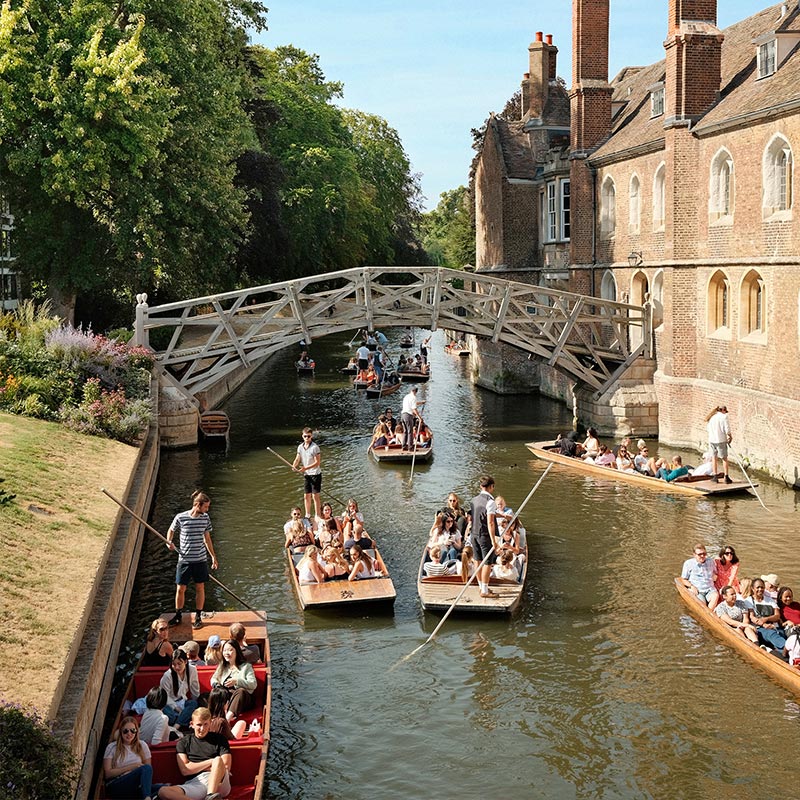 People enjoying a sunny day punting on the River Cam in Cambridge. Several boats are navigating the river, passing under the historic Mathematical Bridge. The scene is lively, with lush greenery and the picturesque architecture of the university buildings lining the riverbank.






