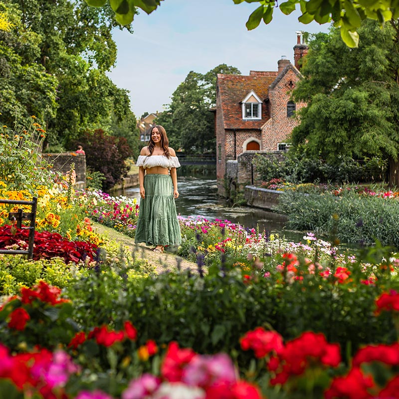 A woman in a white off-the-shoulder top and green skirt stands in a lush, colorful garden filled with blooming flowers. Behind her is a charming red-brick house by a tranquil canal, surrounded by greenery. The scene exudes a picturesque and idyllic atmosphere, showcasing the beauty of the garden and the quaint architecture.






