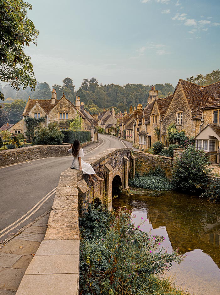 A woman in a white dress sits on the stone parapet of a quaint bridge in Castle Combe, Cotswolds, gazing at the idyllic village and its charming stone cottages. The tranquil river below reflects the greenery and flowers, while the winding road leads through the picturesque scenery.