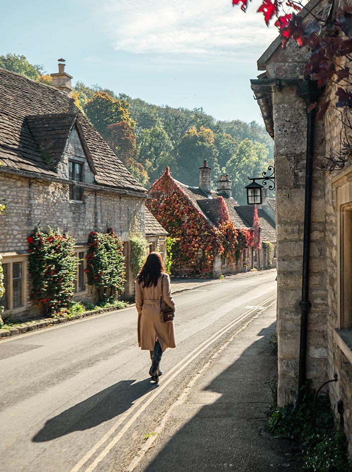 A woman in a beige trench coat walks down a picturesque street in Castle Combe, a charming village in the Cotswolds. The stone houses are adorned with climbing plants and flowers, and the surrounding hills are lush with autumn foliage.