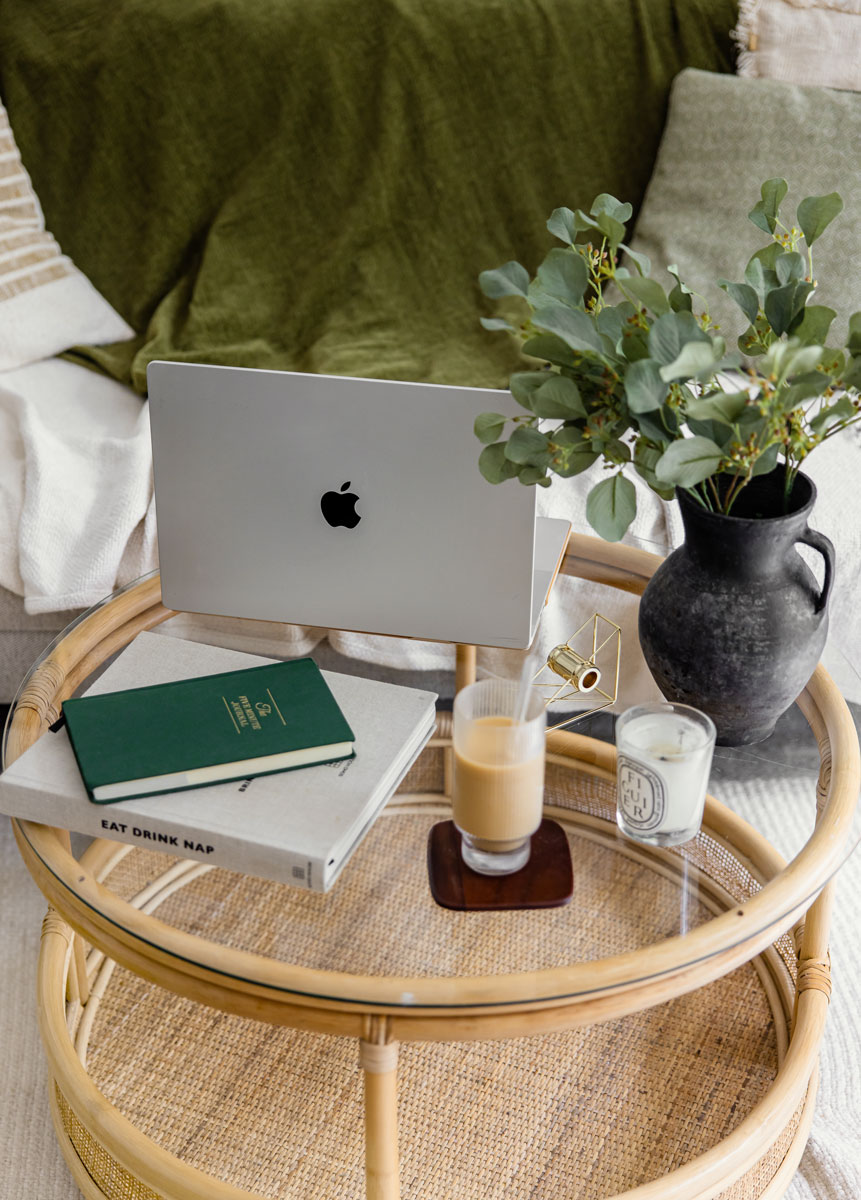 A cozy workspace setup featuring an open MacBook, a green journal, and a book titled "Eat Drink Nap" on a glass-topped wicker coffee table. A vase with greenery, a lit candle, and a glass of iced coffee complete the scene.