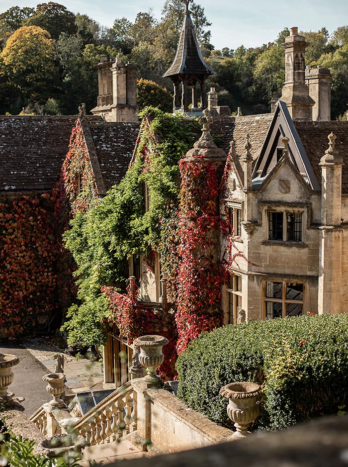 An ornate manor house in the Cotswolds is enveloped by lush green and red ivy. The building's historic architecture features steeply pitched roofs, intricate stonework, and tall chimneys, set against a backdrop of autumn trees. Stone urns and balustrades line the steps leading to the garden below.