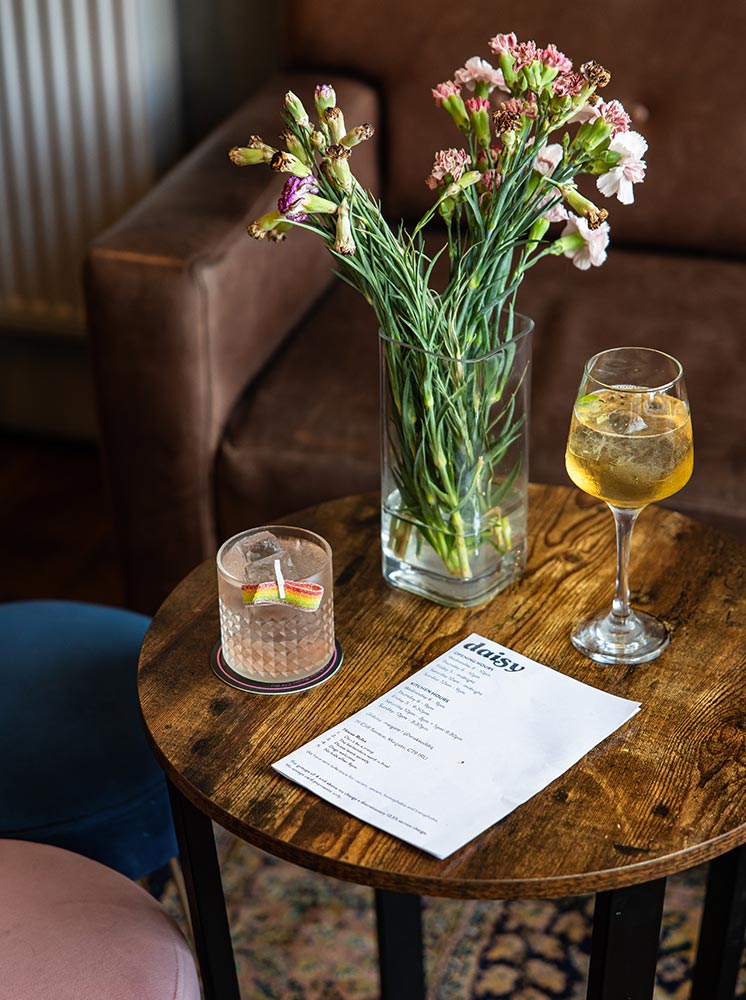 A cozy setting at Daisy Bar, featuring a wooden table with a vase of fresh flowers, a cocktail garnished with a rainbow candy, and a glass of white wine. The table also holds a menu, and comfortable seating is visible in the background, creating a welcoming and relaxed atmosphere.