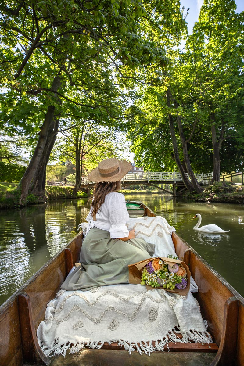 A woman in a white blouse and wide-brimmed straw hat sits in a wooden boat on a serene river, surrounded by lush greenery. A swan glides nearby, and a bouquet of flowers rests beside her. The scene captures a peaceful moment with a quaint bridge visible in the background, reflecting the tranquil beauty of nature.