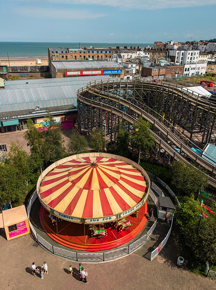 An aerial view of Dreamland amusement park in Margate, featuring a classic carousel with a red and yellow striped canopy in the foreground. The park includes a wooden roller coaster and several buildings with "Margate Amusements" and "Casino Slots" signs. The beach and ocean can be seen in the background, along with a mix of residential and commercial buildings.