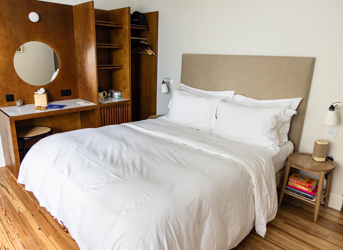 A modern bedroom with a beige upholstered headboard and crisp white bedding. The room features wooden furnishings, including a built-in wardrobe and desk with a round mirror, and a wooden side table with a stack of books. The floor is made of polished wood, and the walls are painted white, creating a clean and cozy atmosphere.






