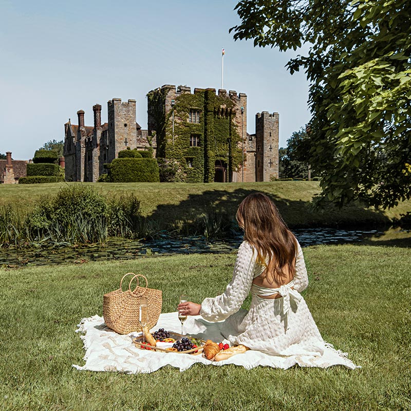 A woman in a white dress sits on a picnic blanket on the lawn, enjoying a picnic in front of the historic Hever Castle. The spread includes grapes, croissants, and a glass of wine. The castle, covered in ivy and surrounded by manicured gardens and a moat, provides a picturesque backdrop under a clear blue sky.