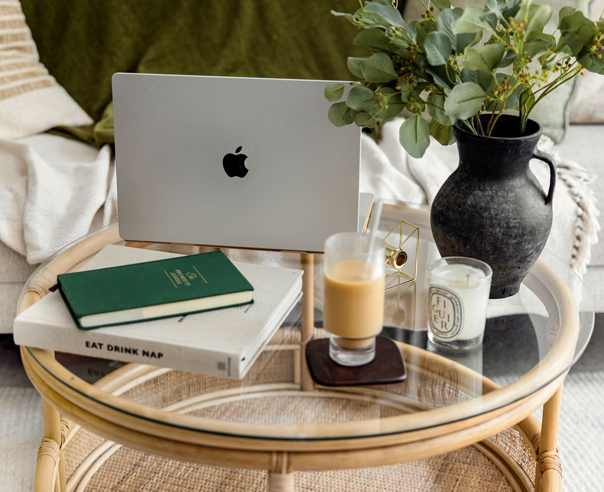 A cozy workspace setup featuring an open MacBook, a green journal, and a book titled "Eat Drink Nap" on a glass-topped wicker coffee table. A vase with greenery, a lit candle, and a glass of iced coffee complete the scene.






